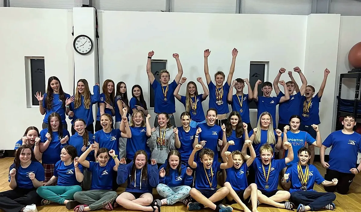 A big group of Academy Swim Team swimmers in blue kit cheering with their arms in the air, many wearing medals