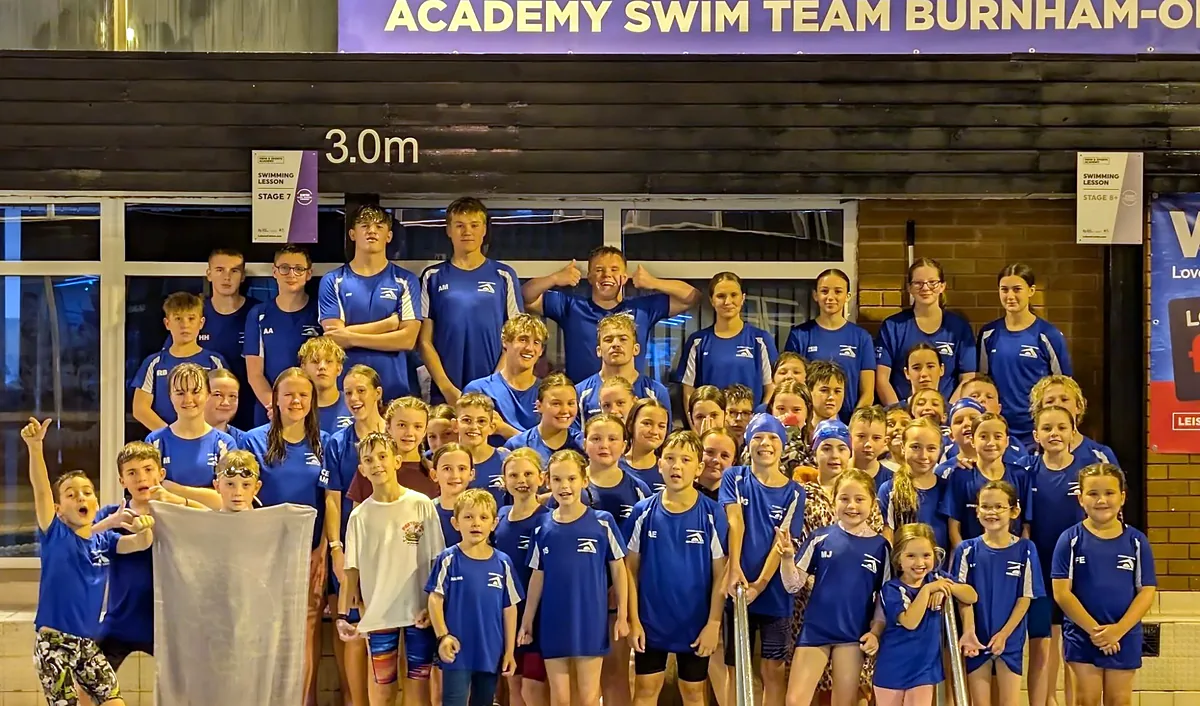 A large group of Academy Swim Team swimmers in blue club kit standing together on the poolside beneath the club banner