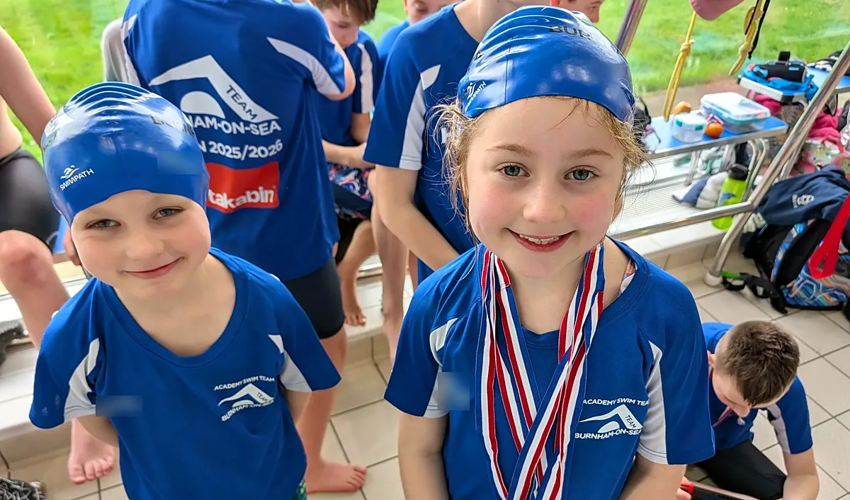 Two young swimmers in blue Academy Swim Team kit and swim caps smiling, one wearing three medals