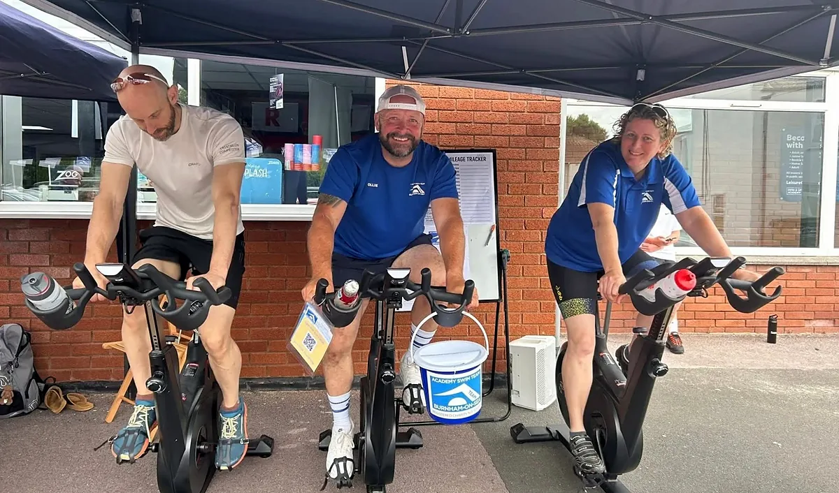 Three people on exercise bikes beside the pool entrance with a club collection bucket, taking part in a fundraising challenge
