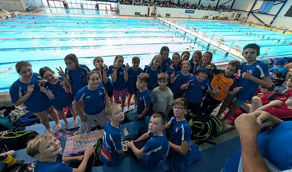 A group of young swimmers in blue club kit smiling beside a large indoor pool