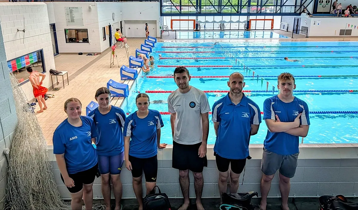 Three young swimmers and three coaches in Academy Swim Team kit standing on the poolside at Burnham Swim & Sports Academy, with the 25 metre pool behind them